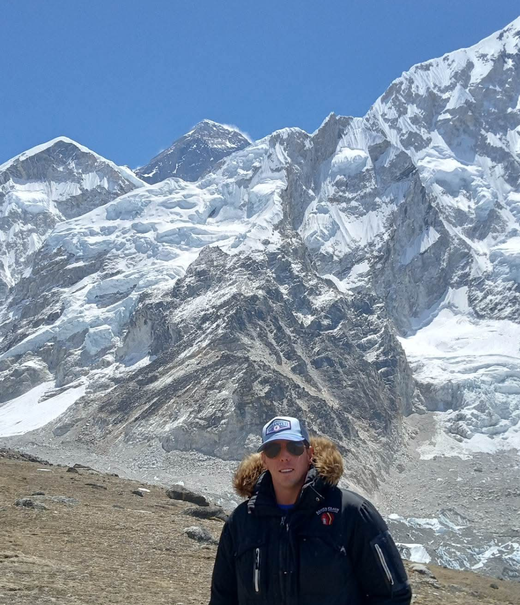 A man wearing a parka, hat, and sunglasses stands in the foreground of a massive, snow-covered mountain range under a clear blue sky.